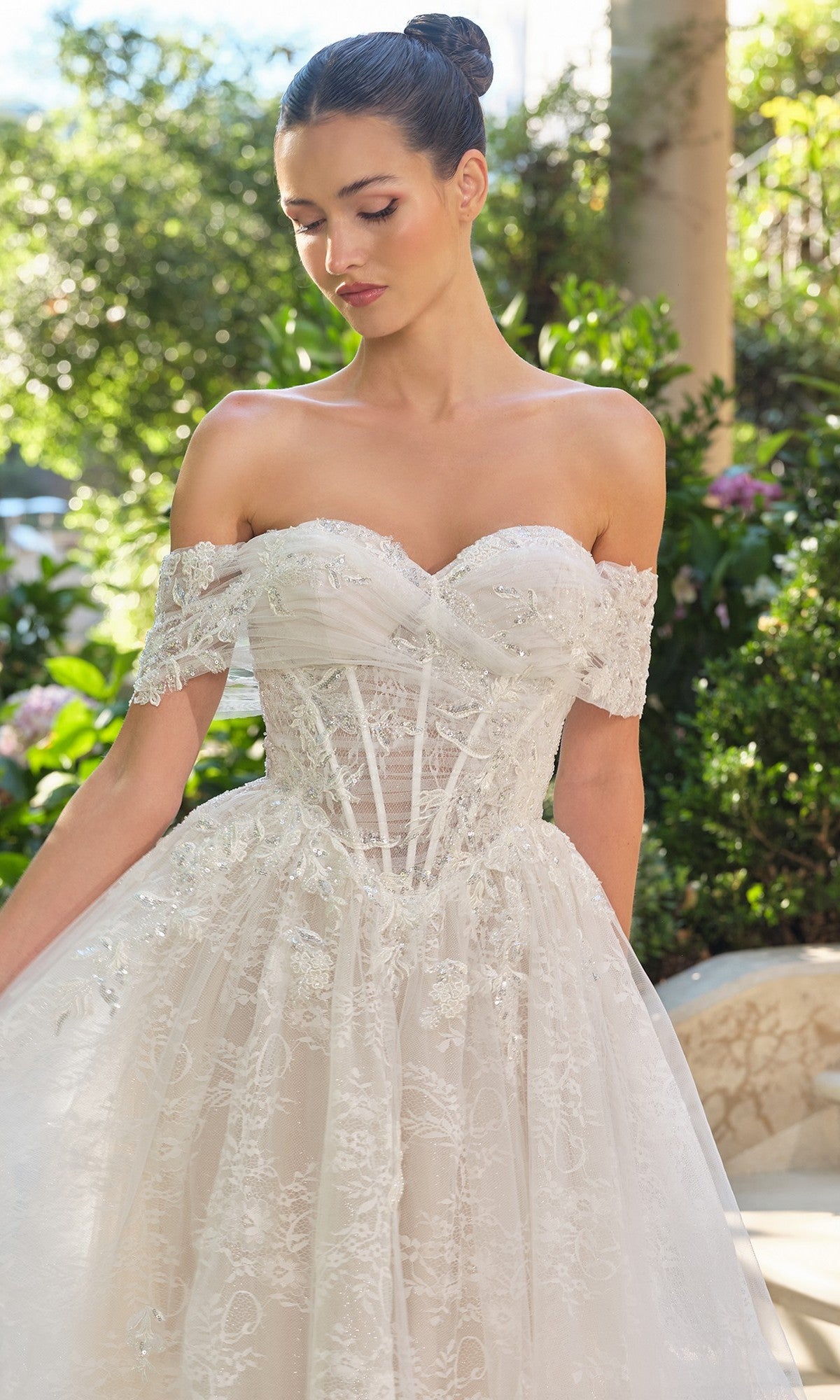 A woman wearing the Long Wedding Dress: Andrea and Leo WL090, featuring an off-the-shoulder corset bodice and lace embellishments, stands outdoors surrounded by lush greenery.